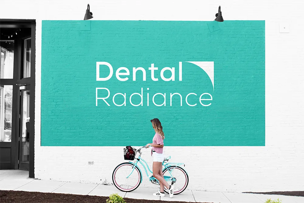 Teal mural sign reading 'Dental Radiance' on a white wall, with a woman in pink and white riding a light blue bicycle in front.