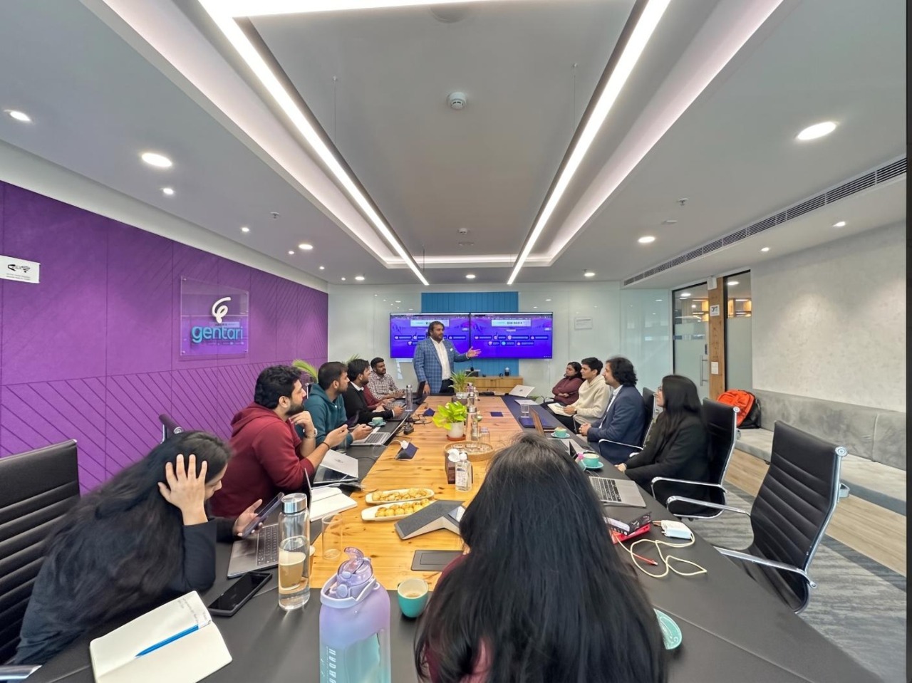 A diverse group of professionals around a long conference table listening to a speaker at the front of the room with a large blue presentation screen behind them.