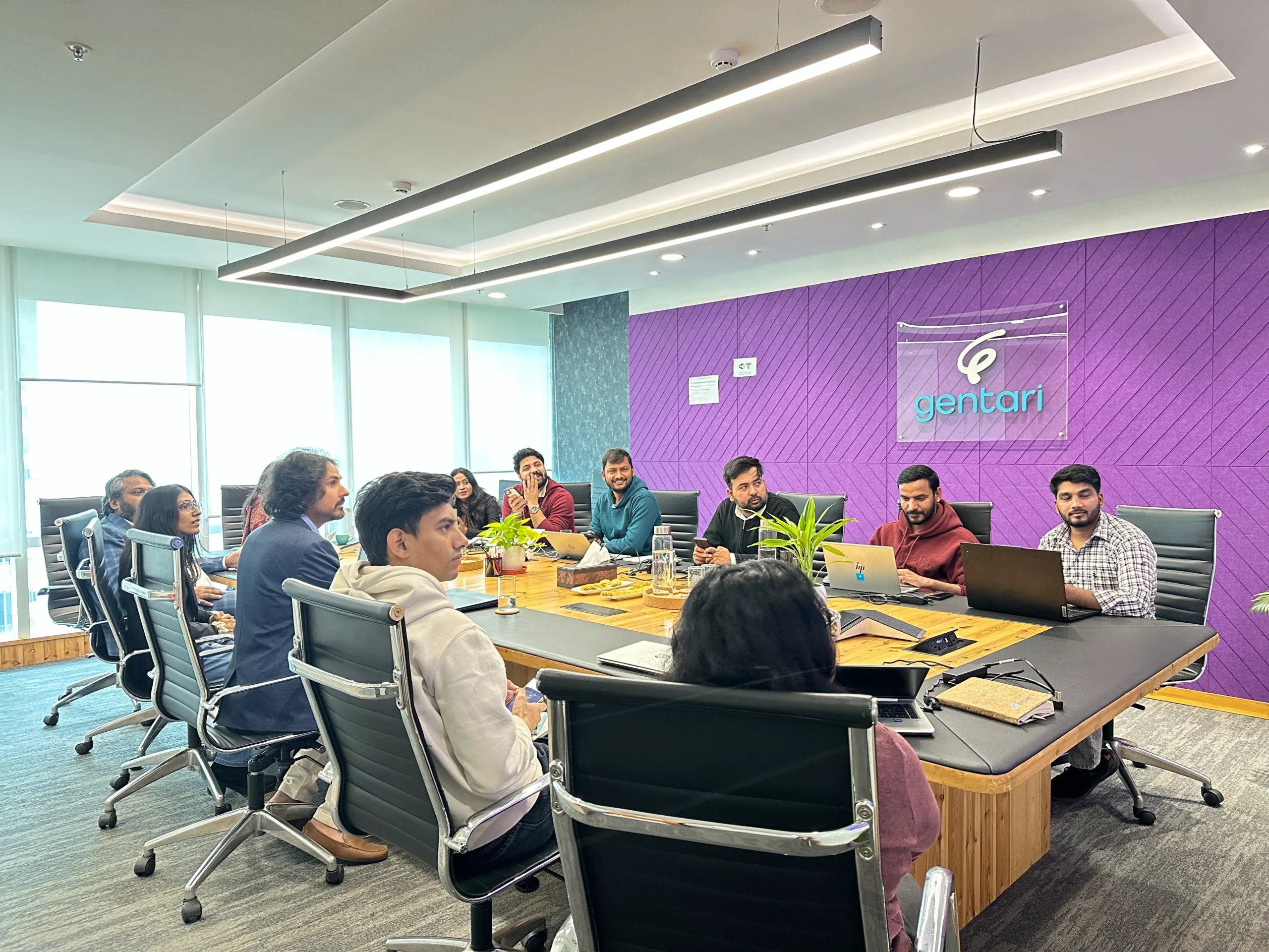 A group of people seated around a long conference table in a modern office, discussing or listening in a meeting, with a purple wall and a transparent panel that reads 'gentari' in the background.