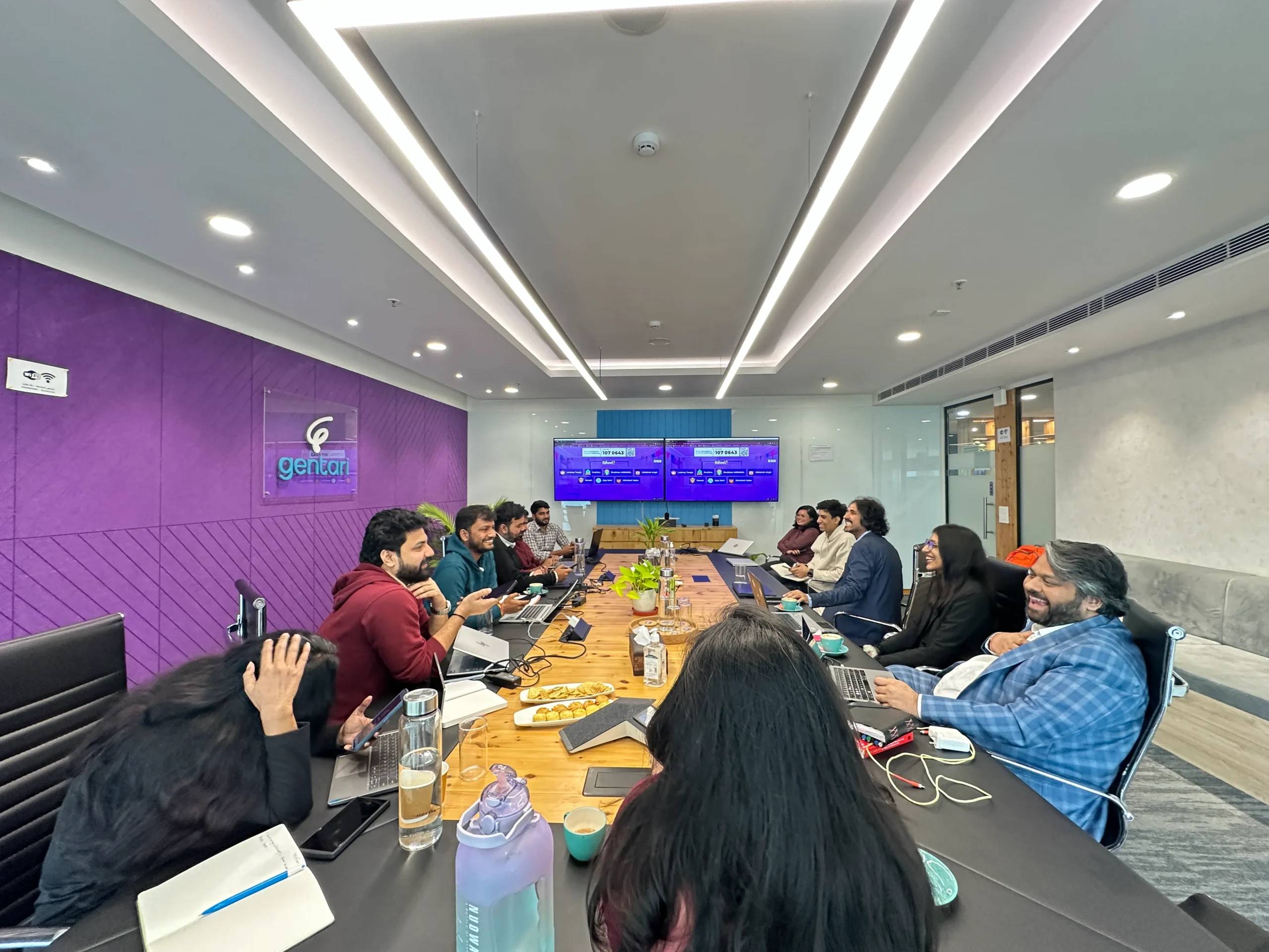 People sit around a long conference table in a bright room with laptops open and snacks on the table, a purple wall in the background.