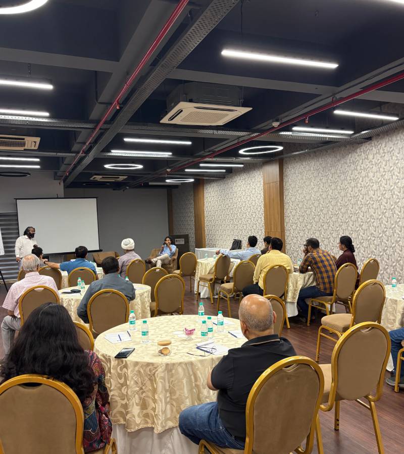 Attendees seated at round tables with water bottles in a conference room, listening to a speaker at the front by a projection screen.