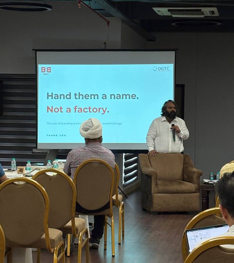 Speaker in white shirt presenting beside a projected slide reading 'Hand them a name. Not a factory.' in a conference room with audience seated at tables.
