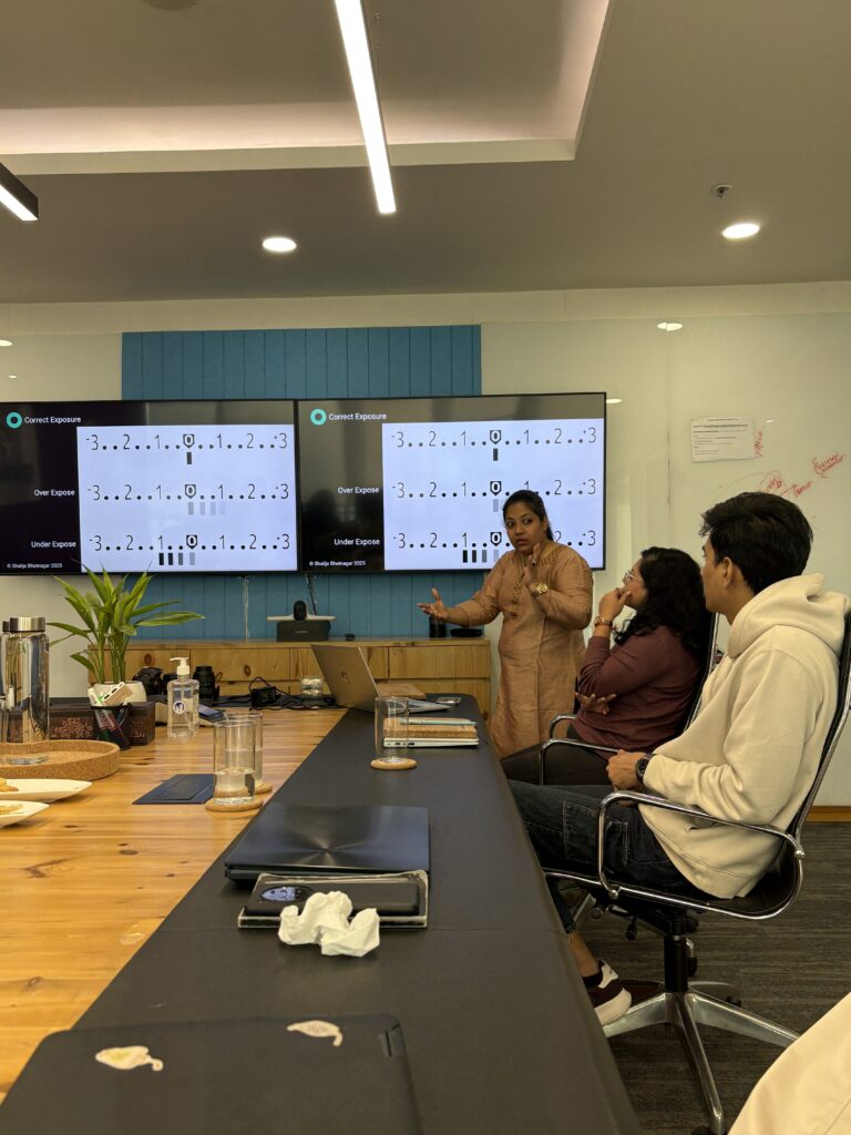 Presenter explaining photography principles to a small group in a meeting room with two large display screens showing a 'Correct Exposure' chart on a blue wall.