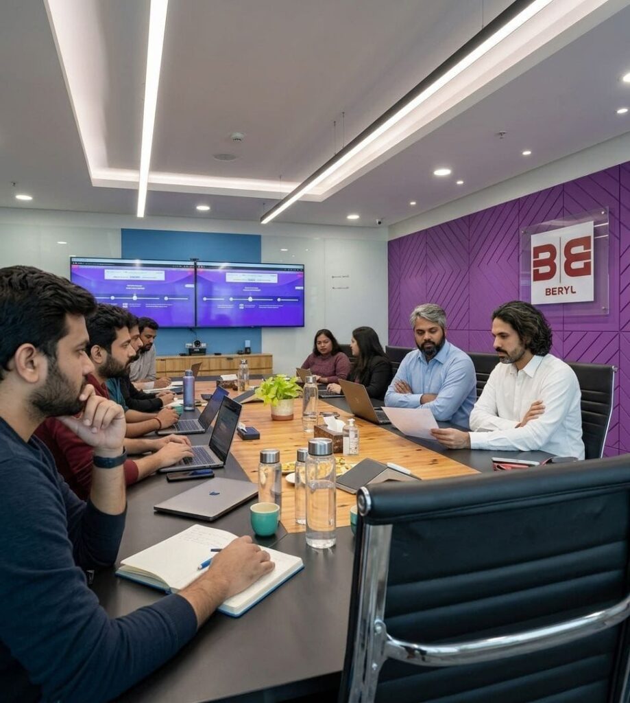 Eight professionals sit around a long conference table with laptops, notebooks, and water bottles, facing two large screens in a glass-walled meeting room with a purple wall and the Beryl logo in the background.