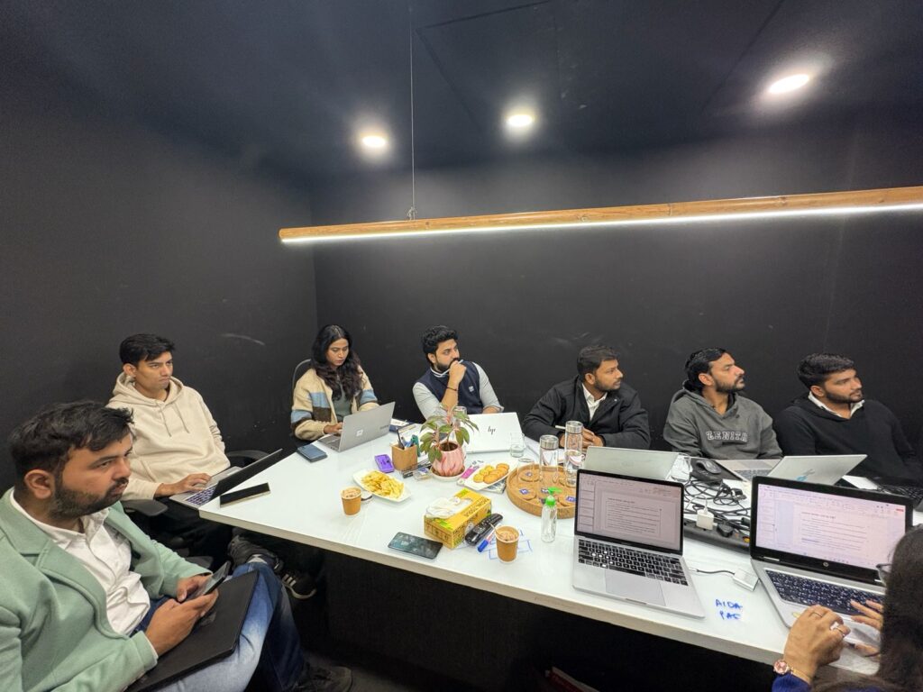Six colleagues sit around a white conference table with laptops, drinks, and snacks in a dimly lit room. plant centerpieces the space as they work.
