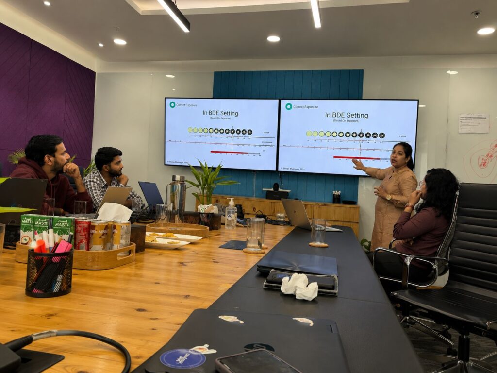 Team in a conference room watching a presentation on two large screens; a woman presenter points to the slide while colleagues listen, laptops open nearby.