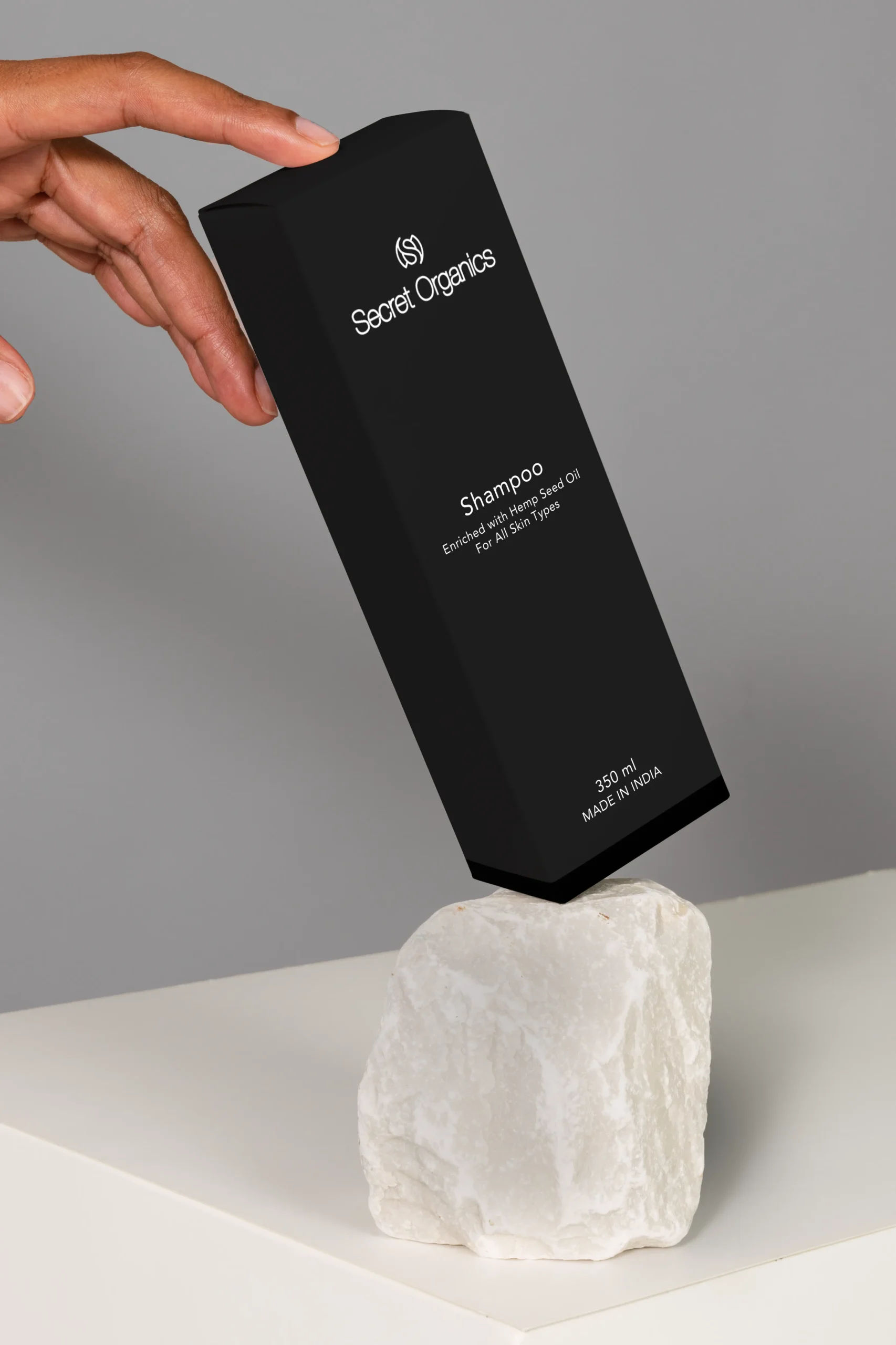 Black Secret Organics shampoo box held by a fingered hand, balanced on a white rock against a gray background, with product text visible: 'Shampoo... Hemp Seed Oil'