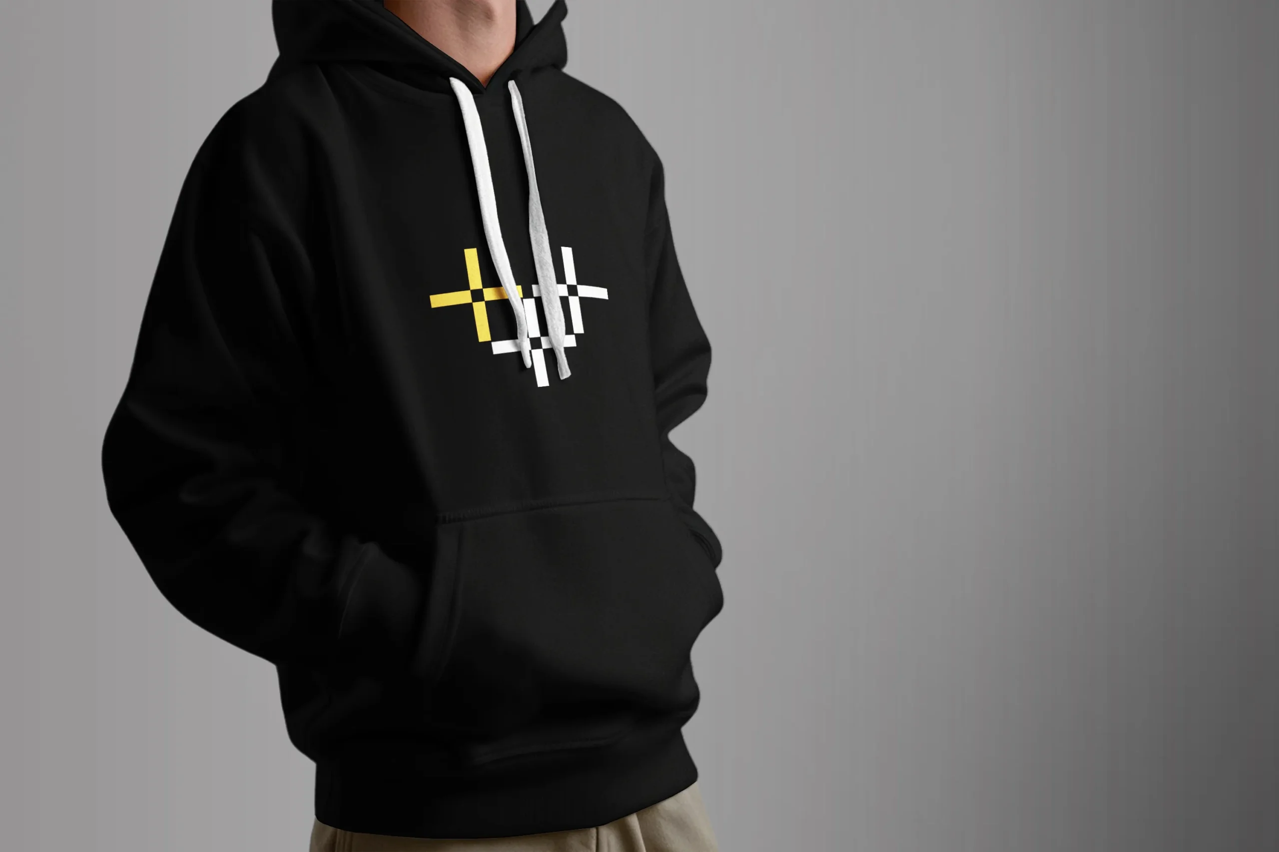 Person wearing a black hoodie with a white-and-yellow geometric cross graphic on the chest, standing against a gray backdrop.