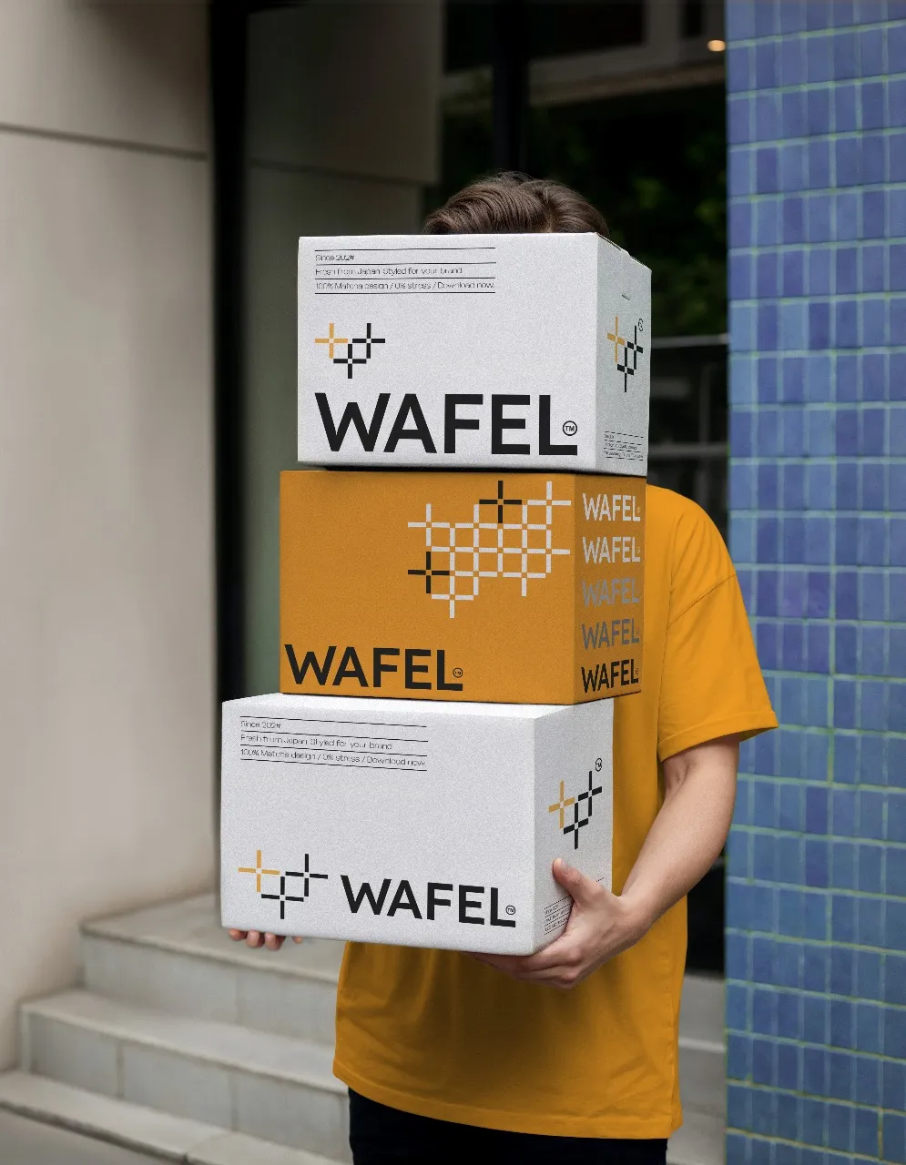 Person in a yellow shirt carrying three stacked WAFEL boxes outdoors.