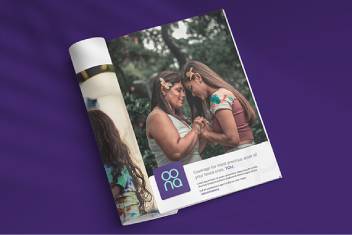 Open magazine spread on a purple surface showing two women in traditional attire touching foreheads, with a logo block and text at the bottom page.