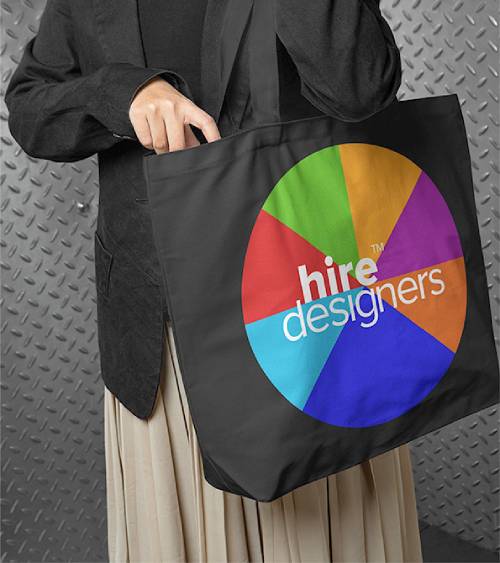 Person in a dark blazer holding a black tote bag featuring a colorful circular logo and the text ‘hire designers’