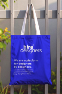 Blue tote bag with white 'hire designers' logo hanging on a metal fence.